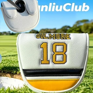 May include: A white golf putter headcover featuring the name GILMORE and the number 18 embroidered in yellow with black outlines. The cover includes a horizontal black stripe and a yellow band at the bottom, with a textured faux leather finish.