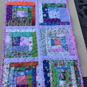 May include: A rectangular table runner featuring a log cabin quilt pattern made from various fabric scraps in shades of pink, green, blue, orange, and white. The blocks are separated by thin strips of light pink fabric with a small floral print.