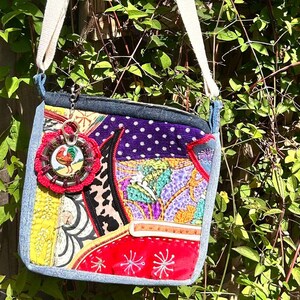 May include: A patchwork crossbody bag with a denim base and a white strap. The bag features a variety of colourful fabric patterns, including polka dots, floral designs, and a red section with embroidered stars. A rooster charm hangs from the bag.