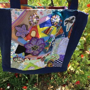 May include: A patchwork tote bag with a denim blue exterior and colourful fabric patches. The bag is adorned with fabric flowers and a dragonfly applique. The bag has blue handles and is set against a backdrop of green foliage and red flowers.