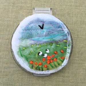 May include: A circular needle minder featuring a needle-felted landscape scene with a blue sky, green rolling hills, and small orange flowers. The design is encased in a clear plastic frame with a white stitched border and a black V-shaped bird.