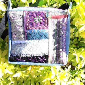 May include: A square crossbody bag featuring a patchwork design of various fabrics including denim, purple, silver, and black textiles. The surface is decorated with intricate embroidery, including floral patterns, cross-stitching, and decorative trim.