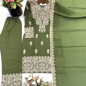 May include: An olive green three-piece outfit consisting of a long-sleeved tunic, trousers, and a sheer dupatta. The tunic features intricate silver embroidery, small floral motifs, and a wide decorative border. The trousers and dupatta match.