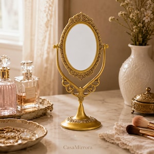 May include: A gold-toned oval tabletop mirror featuring an ornate, embossed frame with intricate scrollwork details. The mirror is supported by a central stand with a decorative base and includes the text CasaMirrora at the bottom center.