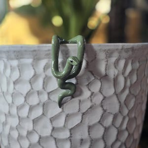 May include: A small, smooth, olive green snake figurine designed to hook over the edge of a container. The snake has a curved body, a defined head with a visible black eye, and a tail that hangs downward against the textured white surface.