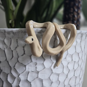 May include: A beige ceramic snake figurine with a smooth, glossy finish and a small black dot for an eye. The snake is coiled and draped over the rim of a white, textured, hammered-finish ceramic planter that features dark grey detailing.