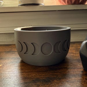 May include: A matte gray cylindrical planter featuring a series of embossed moon phase icons wrapping around the exterior. The design displays the lunar cycle from crescent to full moon, with a smooth finish and a wide, open top opening.
