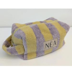 Può includere: Un beauty case rettangolare a righe verticali gialle e lavanda, con manico. Ha una chiusura a zip e una piccola etichetta rettangolare con la scritta "NEA".