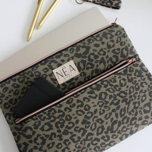 May include: A laptop sleeve with an olive green and black leopard print pattern, featuring a rose gold zipper and a rectangular fabric label with the text NEA bags. A black notebook is tucked into the front zippered pocket of the sleeve.