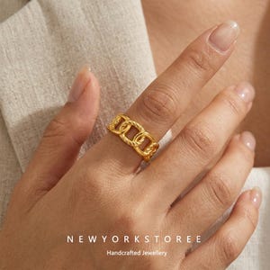 Könnte beinhalten: Ein goldfarbener Ring mit einem sich wiederholenden Kettenglied-Design und einer gedrehten, strukturierten Oberfläche. Das Band besteht aus miteinander verbundenen ovalen Gliedern, die ein durchgehendes Muster um den Finger bilden. Der Text NEWYORKSTOREE Handcrafted Jewellery ist unten sichtbar.