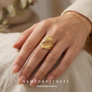 Könnte beinhalten: Ein goldfarbener Ring mit zwei überlappenden Farnblattmotiven in matter Ausführung. Der Ring wird am Ringfinger getragen und ein dünnes goldfarbenes Armband ist am Handgelenk sichtbar. Der Text NEWYORKSTOREE Handcrafted Jewellery ist vorhanden.