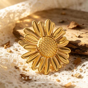 Könnte beinhalten: Eine goldfarbene Metallbrosche in Form einer Gänseblümchenblüte mit einer texturierten mittleren Scheibe und glatten, strahlenden Blütenblättern. Das Stück trägt den Text NEWYORKSTORY Handcrafted Jewellery in Weiß am unteren Rand.