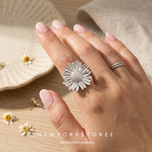 Könnte beinhalten: Ein silberner Ring mit einem großen, detaillierten Gänseblümchen-Design, strukturierten Blütenblättern und einer punktierten Mitte. Der Ring wird am Mittelfinger getragen, ergänzt durch einen dünnen, schlichten Silberring am Ringfinger. Text: NEWYORKSTOREE.
