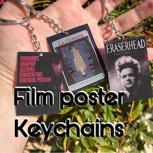 May include: Three rectangular keychains featuring film poster designs. One displays the text Humanist Vampire Seeking Consenting Suicidal Person, another shows the La chimera poster, and the third features the Eraserhead film poster with a portrait.