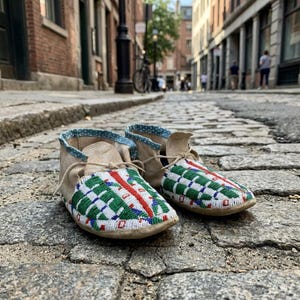 May include: A pair of tan leather moccasins featuring a vibrant geometric beadwork pattern in white, green, blue, and red on the vamp. The footwear includes a light blue patterned fabric trim along the collar and thin leather laces for closure.