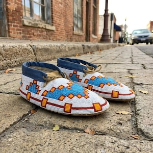 May include: A pair of moccasins featuring a white base with intricate geometric beadwork in shades of blue, orange, and red. The footwear has a blue fabric collar and tan leather accents, with thin laces tied at the top of each shoe.