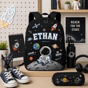 May include: A black backpack featuring a space theme with an astronaut sitting on a moon, planets, stars, and the name ETHAN printed in large white letters. The design includes the text DREAM BIG, EXPLORE, and BE BRAVE in a colorful outer space scene.
