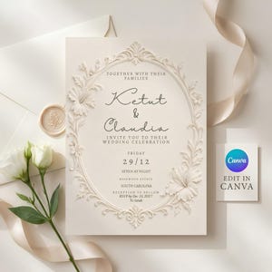 May include: A cream-colored wedding invitation card featuring an embossed oval floral frame with hibiscus flowers. The text reads: Together with their families, Ketut & Claudia, invite you to their wedding celebration, Friday 29/12, seven at night.