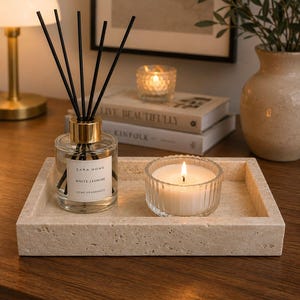 May include: A rectangular beige travertine stone tray holding a clear glass reed diffuser with black sticks and a white label reading Zara Home White Jasmine Home Fragrance, alongside a small ribbed glass candle holder with a lit white wax candle.