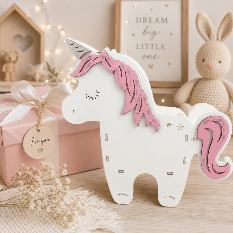 K&ouml;nnte beinhalten: Eine wei&szlig;e Einhornfigur aus Holz mit einem silbernen Glitzerhorn, einer rosa M&auml;hne mit Glitzerakzenten und einem rosa Schweif. Das Einhorn hat geschlossene Augen mit Wimpern und kleine sternf&ouml;rmige Ausschnitte am K&ouml;rper.