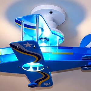 Peut inclure: Un plafonnier en forme d'avion bleu avec des accents blancs et jaunes. Le design comprend une h&eacute;lice blanche, une ampoule centrale et des &eacute;toiles blanches sur les ailes et la queue. Il est fix&eacute; sur une base circulaire blanche.