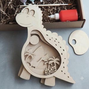 May include: A light-colored wooden dinosaur cutout featuring a layered interior scene with a smaller dinosaur, palm trees, and a sun. The set includes a small wooden paint palette, a paintbrush, and a bottle of red paint inside a cardboard box.