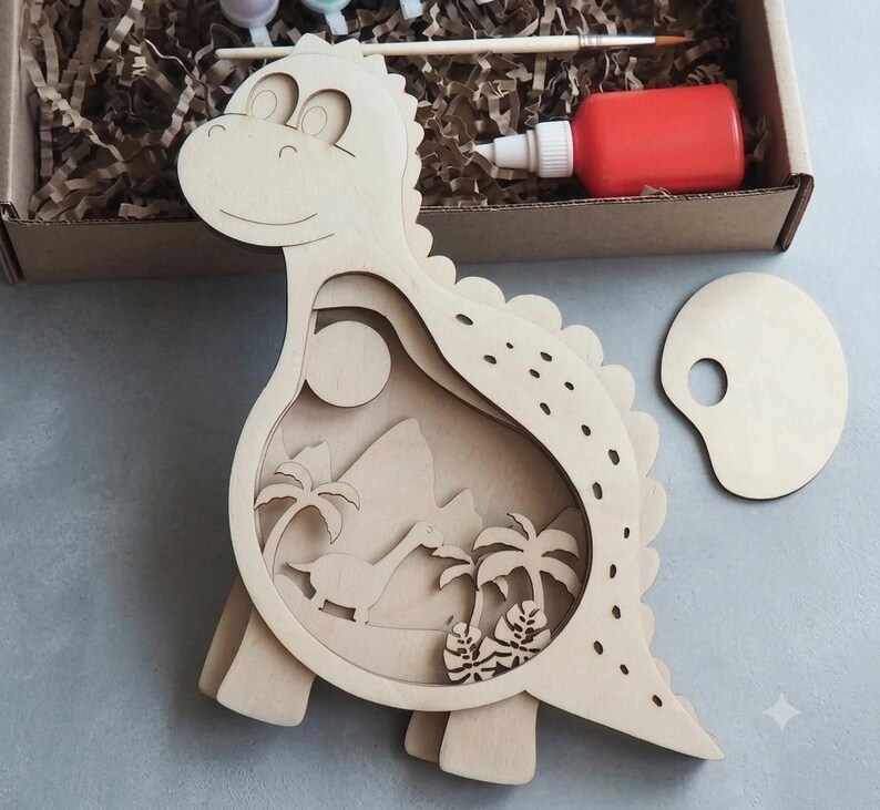 May include: A light-colored wooden dinosaur cutout featuring a layered interior scene with a smaller dinosaur, palm trees, and a sun. The set includes a small wooden paint palette, a paintbrush, and a bottle of red paint inside a cardboard box.