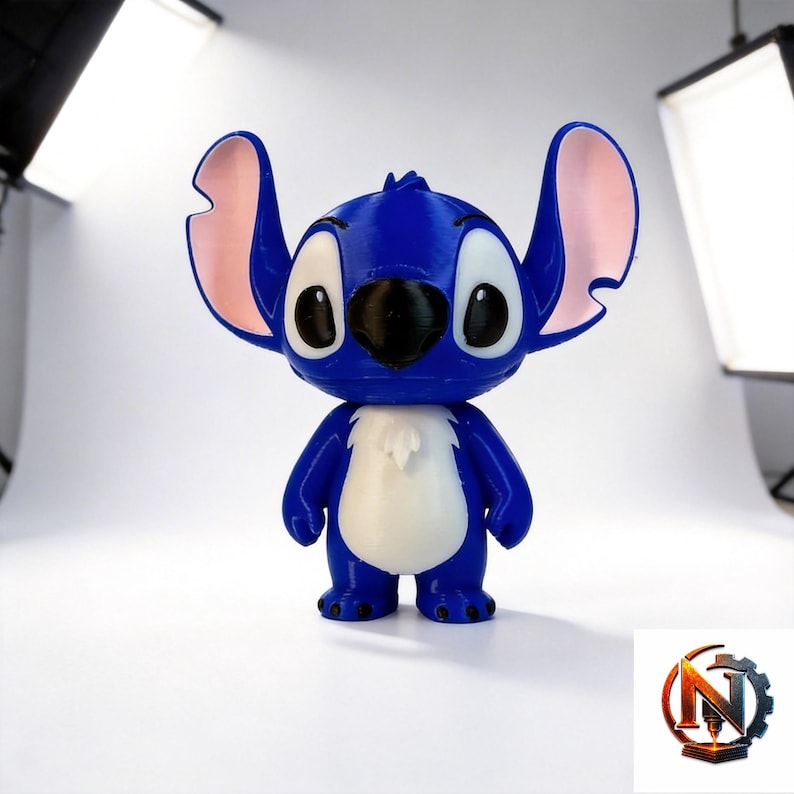 Op de afbeelding: Een blauw-wit beeldje van het personage Stitch met grote oren met roze binnenzijden, grote witte ogen met zwarte pupillen en een zwarte neus. Het figuurtje heeft een glad, afgerond lichaam met een wit vlak op de borst en kleine voetjes.