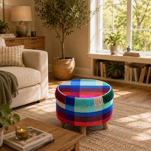 Puede incluir: Un taburete redondo con un vibrante patrón de cuadros multicolor en tonos azul, rojo, verde y amarillo. El borde superior está decorado con flecos a juego y la pieza se apoya sobre cuatro patas cortas de madera de color claro.
