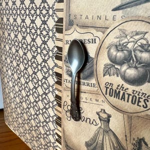 May include: A decorative panel featuring a vintage-style collage with a metal spoon attached. The design includes a black and white geometric pattern, a circular graphic of tomatoes on the vine, a dress illustration, and text reading SHEER, STAINLESS, FRESH, and FARM EGGS.