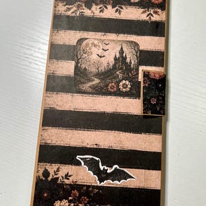 May include: A rectangular card featuring horizontal black and beige stripes, decorated with floral borders, a central rectangular inset showing a castle under a full moon with bats, and a separate bat cutout positioned near the bottom edge.