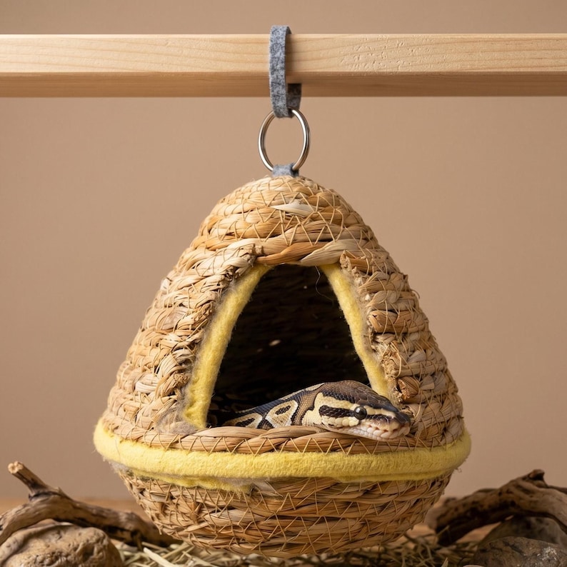May include: A woven, teardrop-shaped hanging pet hideaway made of natural tan fibers with a yellow felt trim around the triangular entrance. A patterned snake is resting inside the structure, which is suspended by a metal ring and grey felt loop.