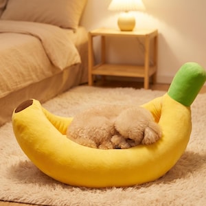 May include: A plush banana-shaped pet bed featuring a bright yellow curved body, a green stem at one end, and a brown tip at the other. A small, light-colored, curly-haired dog is curled up and sleeping inside the soft, cushioned center of the bed.