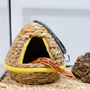May include: A woven straw dome-shaped pet hideout with a triangular opening trimmed in bright yellow fabric. A grey felt strap with a metal ring is attached to the top. An orange and white patterned corn snake is emerging from the entrance.