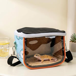 May include: A blue pet carrier with a clear front and a black mesh top. The carrier features a cartoon bear design and an orange trim. Inside, a leopard gecko rests on a checkered surface. Includes a black shoulder strap.