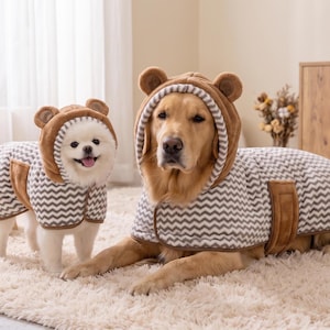 Può includere: Due cani indossano cappottini coordinati per animali domestici con motivo chevron marrone e bianco e cappucci con piccole orecchie da orso. I cappotti presentano bordi marroni e un design avvolgente con una chiusura a fascia marrone laterale.