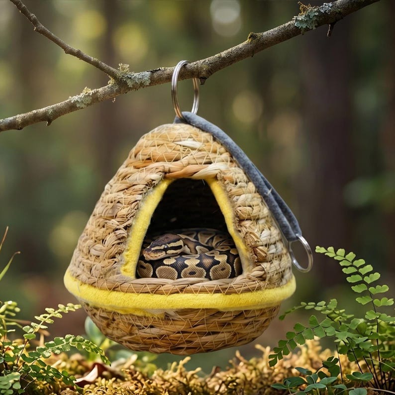 May include: A woven, dome-shaped hanging hideout with a yellow felt trim and a metal ring attachment. Inside the triangular opening, a brown and black patterned ball python is coiled, resting within the textured, natural fiber structure.