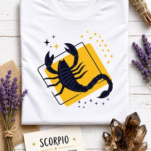 May include: A white t-shirt featuring a graphic of a black scorpion centered on a yellow square. The design includes small yellow dots and dark blue stars. Below the shirt, a card displays the text: SCORPIO, STRENGTH & INTUITION, 13 NOV - 21 NOV.
