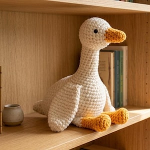 Op de afbeelding: Een handgehaakt ganzenknuffel met een crèmekleurig lichaam, een textuur van garen, een feloranje snavel en bijpassende oranje voeten. Het knuffeldier heeft kleine zwarte kraaloogjes en bevindt zich in een zittende houding.
