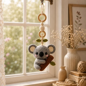 May include: A handcrafted crochet koala baby mobile. The grey koala has a black nose and pink cheeks, clutching a brown branch. The mobile features wooden rings and beads, with green leaf accents. It is suspended from a metal hook.