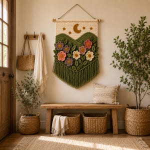 May include: A rectangular crochet wall hanging featuring a cream-colored top section with a brown crescent moon and stars, a green mountain-shaped middle section with various colorful crocheted flowers, and a long green fringe at the bottom.