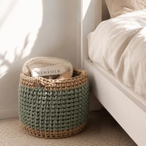May include: A round, hand-crocheted storage basket with a two-tone design. The lower section is sage green, and the upper rim is a natural beige. The basket contains a folded cream-colored blanket and a magazine with the word "KINFOLK" on the cover.