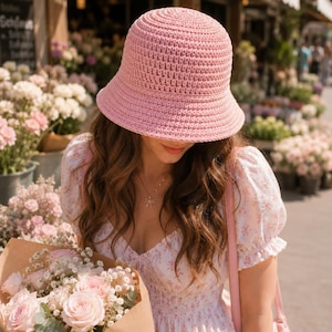 Könnte beinhalten: Ein hellrosa gehäkelter Glockenhut mit breiter Krempe. Die Person trägt ein geblümtes Kleid und hält einen Strauß rosa Rosen. Der Hintergrund zeigt ein Blumengeschäft mit verschiedenen Blumen.