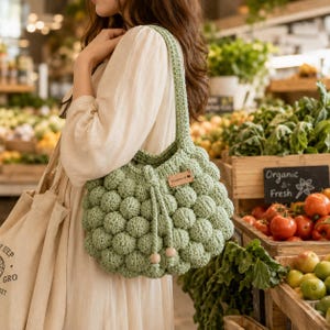 May include: A light green, handmade crochet bag with a unique textured bubble design. The bag has a drawstring closure and wooden bead accents. A small, rectangular tag with the word "Handmade" is attached. The bag is carried in a market setting.