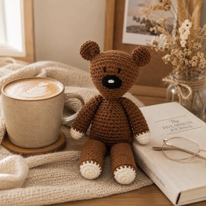 May include: A handmade brown crochet teddy bear with white accents on the feet and a black nose. The bear sits near a cup of coffee and a book titled "The Five Minute Journal". The scene is set on a neutral-toned blanket.
