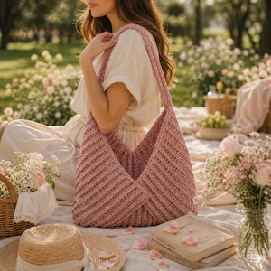 May include: A large, dusty rose-colored crochet tote bag with a wide shoulder strap. The bag has a geometric design and is shown outdoors with flowers and a straw hat, suggesting a picnic setting.