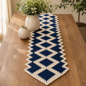 May include: A long, rectangular crochet table runner featuring a repeating geometric diamond pattern in deep navy blue and cream yarn. The runner has a scalloped edge and is laid flat, showcasing the intricate interlocking stitch work throughout.