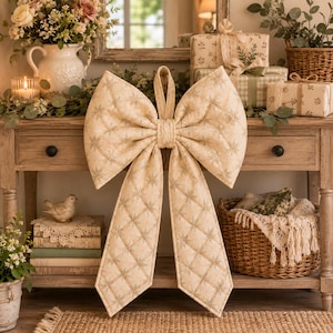 May include: A large, quilted fabric bow in a light beige color featuring a repeating gold metallic snowflake pattern. The bow has two wide, structured loops at the top and two long, pointed tails hanging down, with a central knot detail.