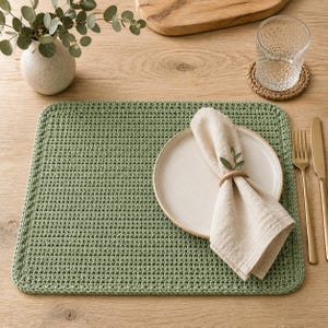 May include: A rectangular sage green crocheted placemat featuring a textured stitch pattern. A round off-white ceramic plate sits on the placemat, topped with a folded beige linen napkin held by a simple wooden ring and a small green leaf sprig.