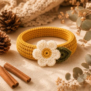 May include: A circular mustard yellow crochet headband featuring a central white flower applique with a yellow center and a single dark green leaf attached to the side, crafted with a dense, textured stitch pattern throughout the band.